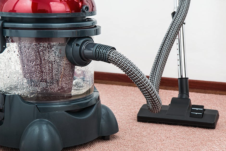 Close-up image of a portable vacuum cleaner with a transparent dust container filled with debris, connected to a flexible hose attached to a vacuum head resting on a pink carpet. The vacuum cleaner is