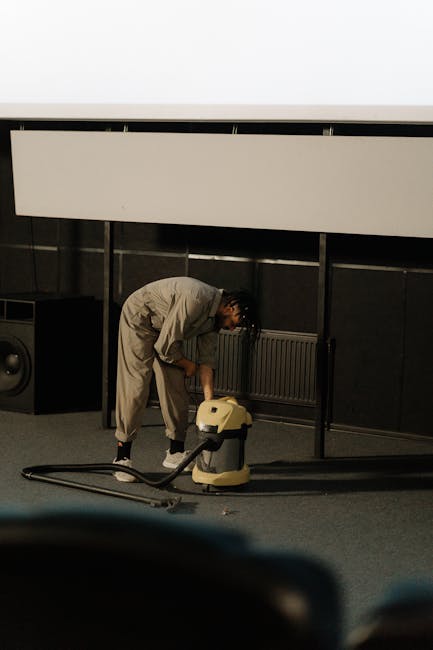 Close-up image of a portable vacuum cleaner with a transparent dust container filled with debris, connected to a flexible hose attached to a vacuum head resting on a pink carpet. The vacuum cleaner is