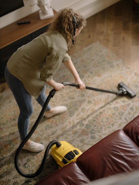A person engaged in surface cleaning within a living room, using a yellow vacuum cleaner with a long hose and nozzle attachment to deep clean a patterned carpet. The individual is dressed casually in 